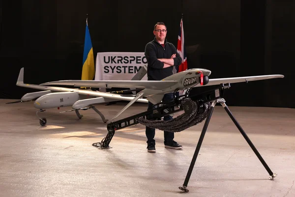 Un homme debout près de deux drones dans un intérieur, sous une bannière UKRSPEC SYSTEMS et des drapeaux nationaux.