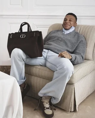 Kylian Mbappé, égérie de la campagne, porte un sweat Dior gris avec un sac à main sur les genoux.