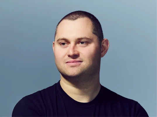Mark Gurman, rédacteur en chef chez Bloomberg, photographié en gros plan avec un t-shirt noir sur fond bleu clair.