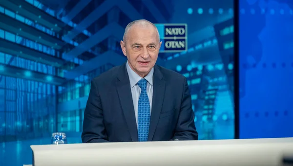 Homme en costume s'adressant à la presse depuis une tribune officielle avec le logo de l'OTAN visible en arrière-plan.