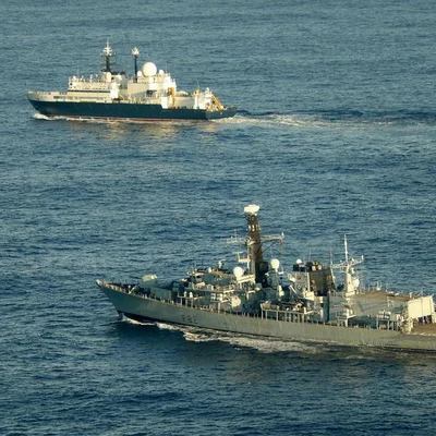 Deux navires militaires sur l'eau, dont un navire de guerre gris au premier plan et un bâtiment plus important en arrière-plan.