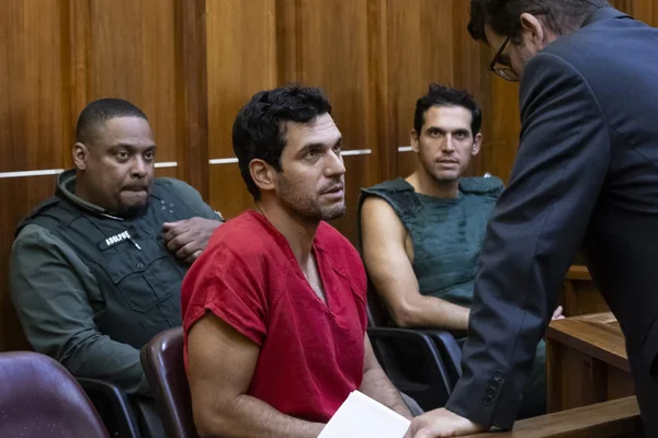 Scène de tribunal avec un homme en tenue de prison rouge, un officier et un homme en costume penché sur une table.