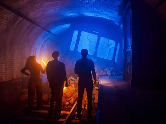 Silhouettes sur les rails dans un tunnel éclairé en bleu devant une voiture de train abîmée lors du Live Cinema.