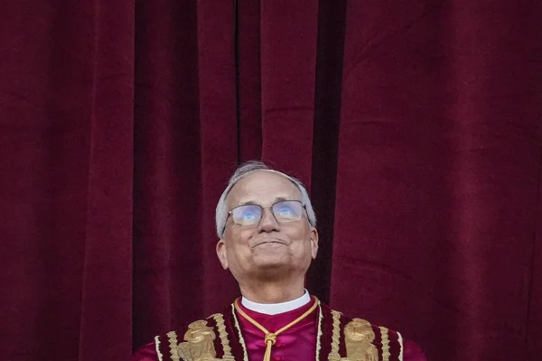 Une personne en vêtements religieux ornés de détails bordeaux et or, debout devant un rideau.