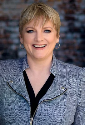 Alison Arngrim portant une veste bleue et blanche à motifs, souriante devant un mur de briques.