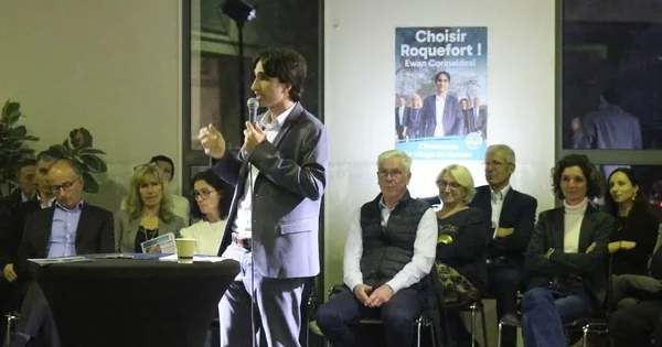 Ewan Corinaldesi saluant la foule devant son panneau de campagne la nuit.