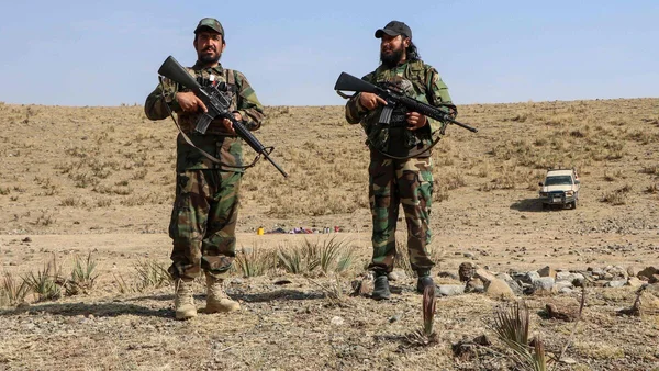 Deux hommes en uniforme militaire tiennent des fusils dans un paysage désertique, avec un véhicule à l'arrière-plan.