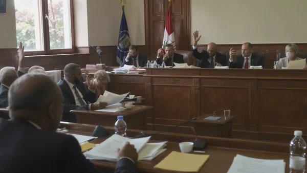 Séance législative dans une salle boisée avec des députés levant la main pour voter, documents et drapeaux visibles.