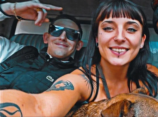 Vue de l'intérieur d'un véhicule montrant une femme tatouée et un homme en lunettes de soleil à ses côtés.