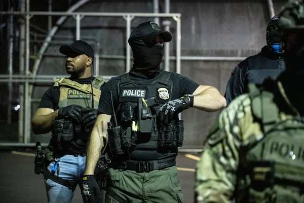 Trois policiers en tenue d'intervention, dont un porte un masque facial, dans un décor industriel.