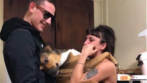Un homme et une femme tenant un chien, debout dans une intérieur semblant être une cuisine.