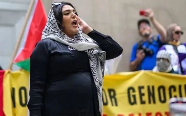 Nerdeen Kiswani portant un keffiyeh lors d'une manifestation avec des banderoles jaunes visibles à l'arrière-plan.