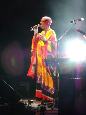 La chanteuse Desireless, coiffée de cheveux blancs et portant une robe colorée, chante sur scène.