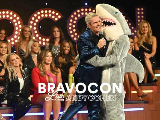Andy Cohen en costume bleu serrant une personne en costume de requin sur scène lors du BravoCon Live.