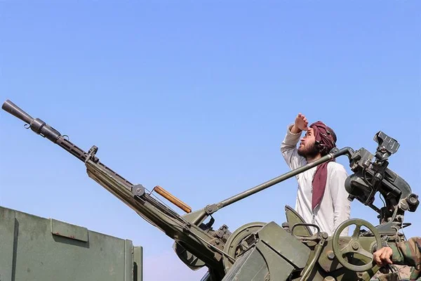 Un homme portant un turban rouge manipule une mitrailleuse montée sur un véhicule militaire sous un ciel bleu.