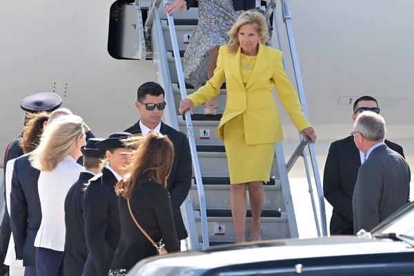 Jill Biden en costume jaune descendant d'un avion, escortée par des agents de sécurité sur le tarmac.