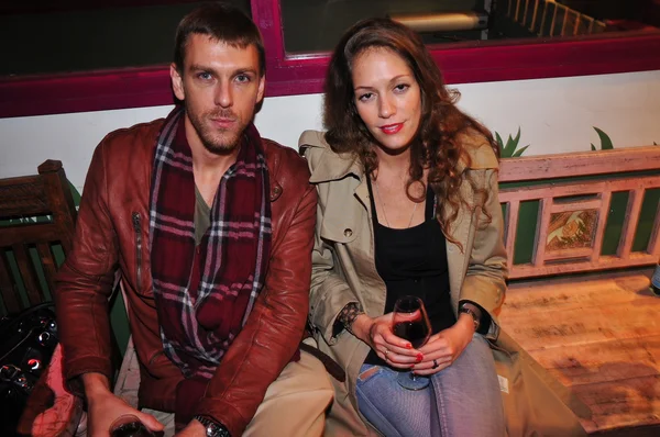 Un homme en veste de cuir rouge et une femme en trench-coat beige tenant un verre de vin sur un banc.