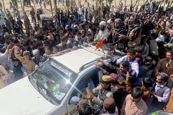 Une grande foule entoure un véhicule blanc surmonté d'une fleur rouge et d'hommes armés sous un ciel dégagé.