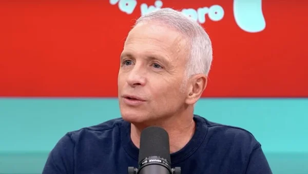 Samuel Etienne parlant au micro dans un studio télévisé aux couleurs rouge et turquoise.