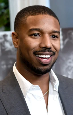 Michael B. Jordan en costume gris et chemise blanche, affichant un sourire confiant.