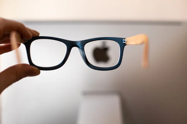 Des lunettes à monture bleue tenues en main, le logo Apple apparaissant en flou derrière.