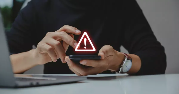 Une main tenant un téléphone portable signalé par un triangle de danger rouge, posé sur une table devant un ordinateur.