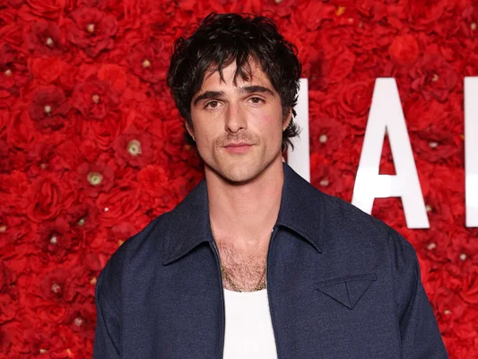 Jacob Elordi en veste bleue devant un fond floral rouge éclatant.