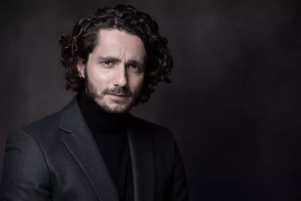Guillaume Pley en veste sombre et col roulé noir, sur un fond sombre texturé.