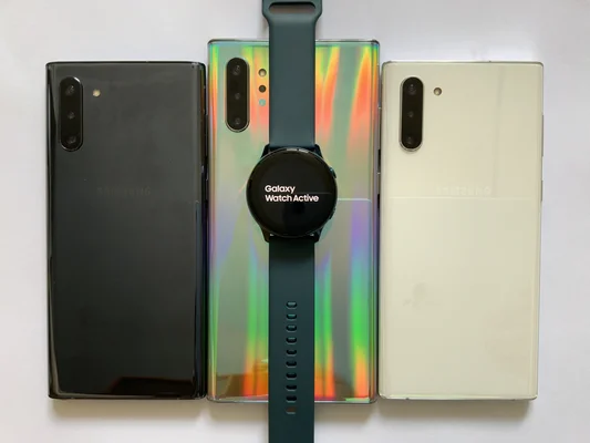 Trois smartphones Samsung alignés avec une Galaxy Watch Active posée sur le téléphone central sur fond blanc.