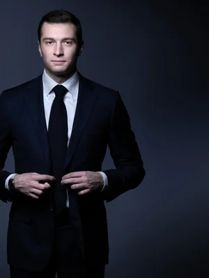 Jordan Bardella en costume bleu nuit ajustant sa veste sur fond sombre.