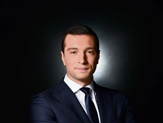 Jordan Bardella en costume bleu marine et cravate foncée sur fond sombre.