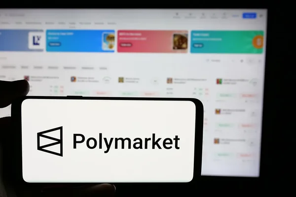 Un smartphone affichant le logo de Polymarket tenu par une main devant un écran d'ordinateur.