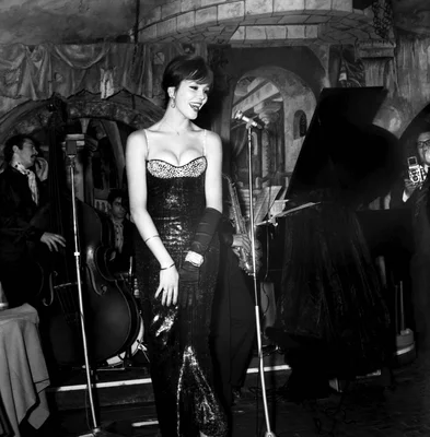 Amanda Lear sur scène dans les années 60, robe scintillante et micro, entourée de musiciens.