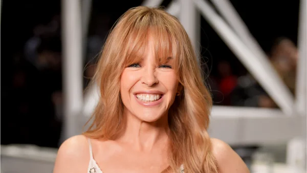 Kylie Minogue en gros plan, arborant des gants rouges et un rouge à lèvres assorti, dans une pose élégante près d'une fenêtre.