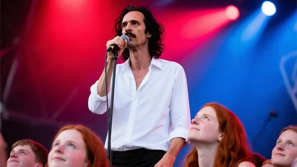 Nick Cave sur scène au Flow Festival, micro à la main, chemise blanche, cheveux ébouriffés, projecteurs rouges et bleus, public en contrebas captivé, ambiance rock intense