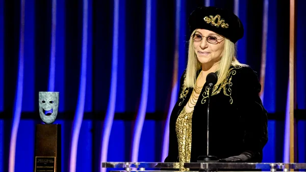 Barbra Streisand au pupitre, lunettes, béret et veste noirs à détails dorés, trophée masque bleu à côté.