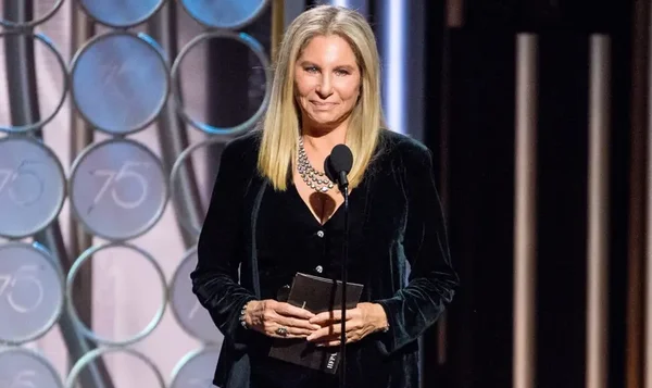 Barbra Streisand au pupitre, blazer velours noir et collier argenté, dossier noir en main.