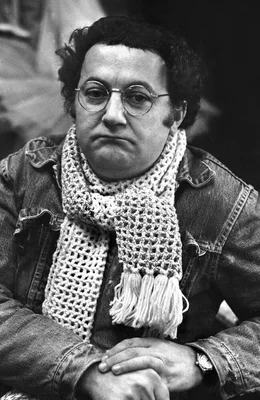 Portrait de Coluche en veste en jean et écharpe crochetée.