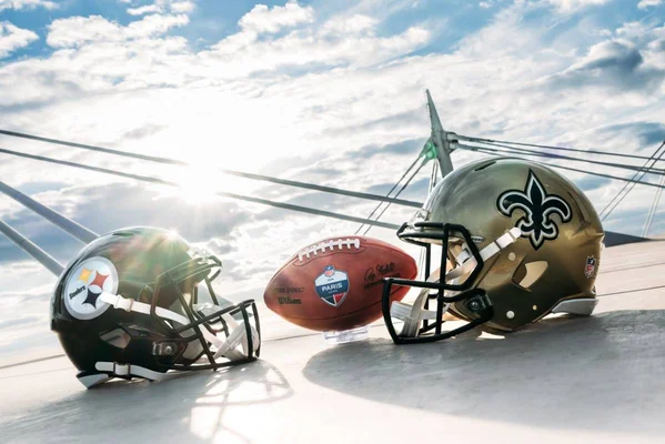 Ballon officiel NFL Paris entre les casques des Steelers et des Saints, avec un pont en arrière-plan.