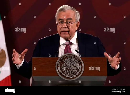 Le Gouverneur de l'État de Sinaloa, Ruben Rocha Moya, aux cheveux gris et portant des lunettes, s'exprimant avec des gestes des mains lors d'une conférence de presse devant le président du Mexique.
