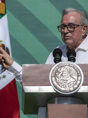 Ruben Rocha Moya, portant des lunettes et une chemise blanche, s'exprimant à un podium arborant le sceau du Mexique, avec le drapeau national à côté.