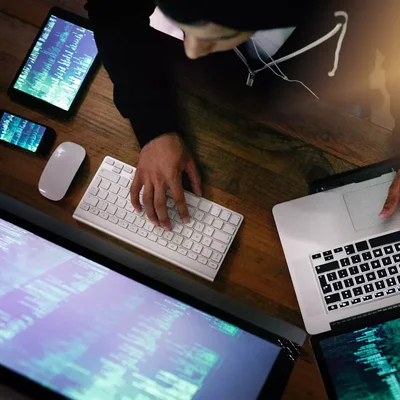 Personne en sweat à capuche noir travaillant devant plusieurs écrans affichant du code, évoquant un environnement de programmation ou de piratage.