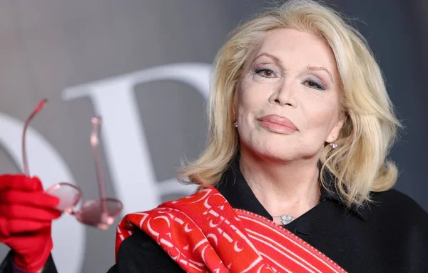 Amanda Lear en manteau noir et écharpe rouge, tenant des lunettes roses.
