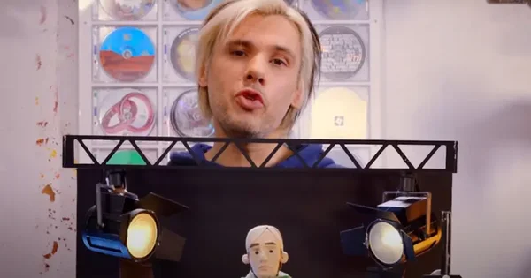 Orelsan derrière une scène miniature avec projecteurs et marionnette, entouré de CD.