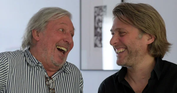 Cookie et Tom Dingler, père et fils, rient ensemble devant un mur blanc.