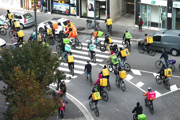 Groupe de livreurs à vélo et scooters des plateformes numériques traversant un carrefour en ville.