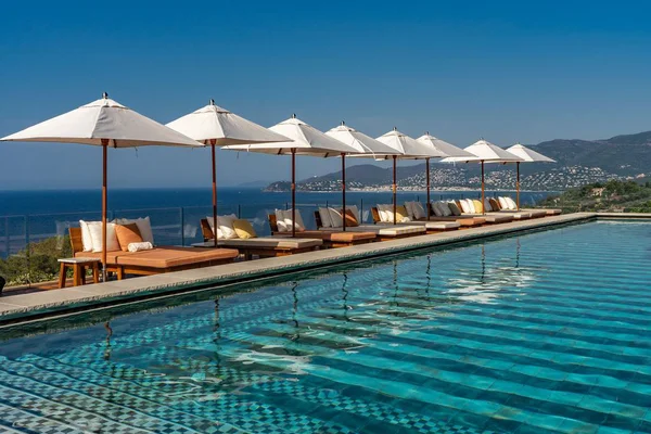 Parasols blancs et chaises longues en bois au bord d'une piscine avec vue sur la côte et les montagnes.
