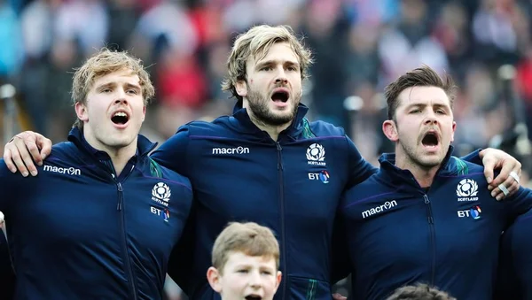 Trois joueurs écossais, en tenue d'équipe, chantant Flower of Scotland avec émotion.