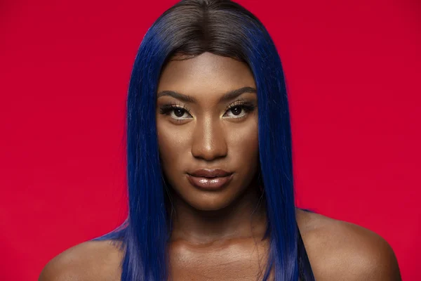 Aya Nakamura, cheveux bleus, en portrait studio sur fond rouge.