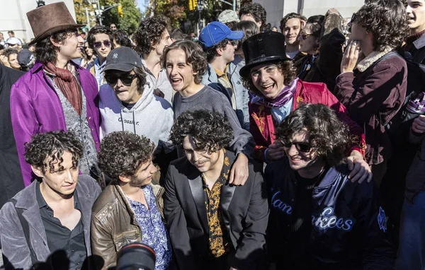 Groupe de jeunes sosies de Timothée Chalamet en tenues vintage et hauts-de-forme, souriant et interagissant en extérieur.
